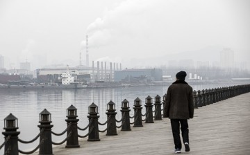 A man strolls along a waterfront boardwalk facing factories and a shipbuilding yard in Dalian, China.