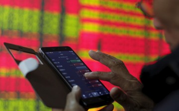 An investor looks at stocks on his mobile phone, in front of an electronic screen showing stock information at a brokerage house in Hangzhou, Zhejiang Province, April 10, 2015. 