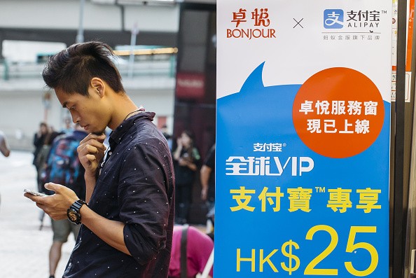 A man with a smartphone stands beside an advertisement for Alipay in a Hong Kong store.