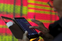 An investor looks at stocks on his mobile phone, in front of an electronic screen showing stock information at a brokerage house in Hangzhou, Zhejiang Province, April 10, 2015. 